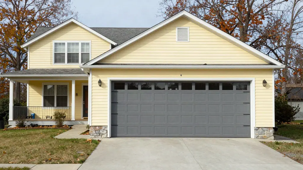 Closed gray sectional garage door with a row of top windows on a pale yellow two-story house.